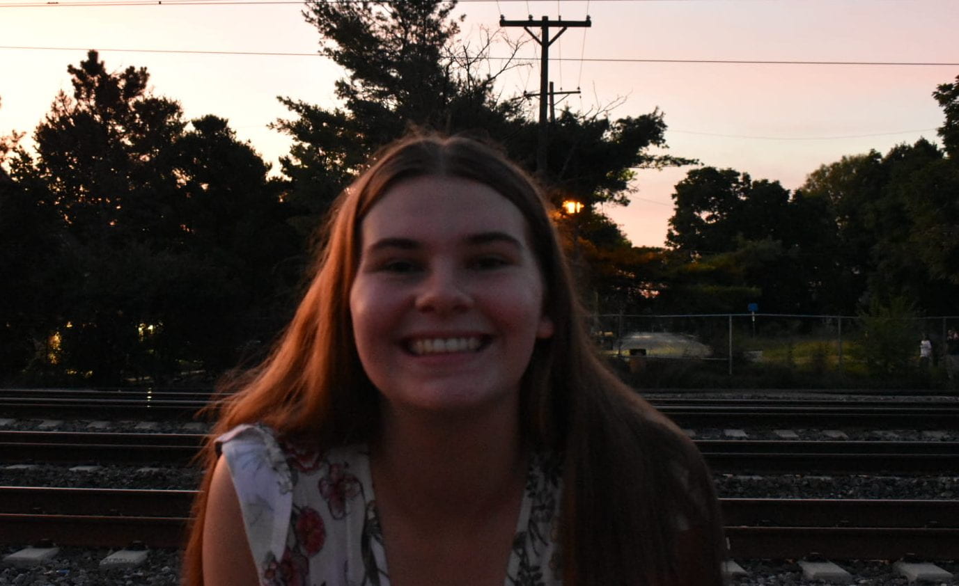 a girl stands crouched forward smiling at the camera in front of train tracks