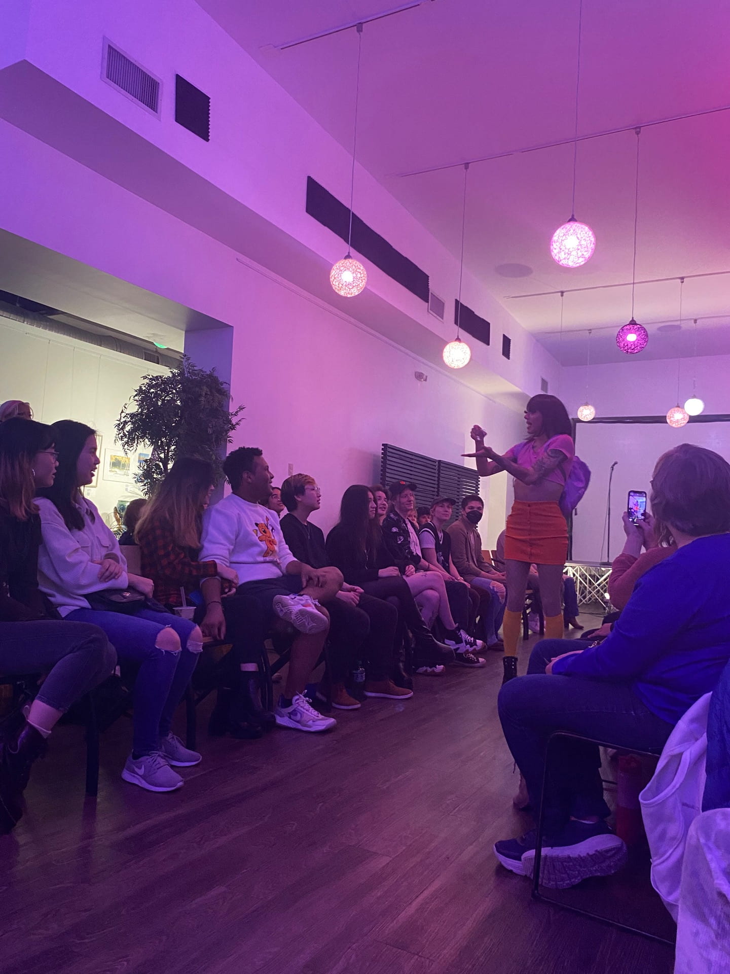 drag queen in an orange skirt stands I purple lit room interacting with seated crowd