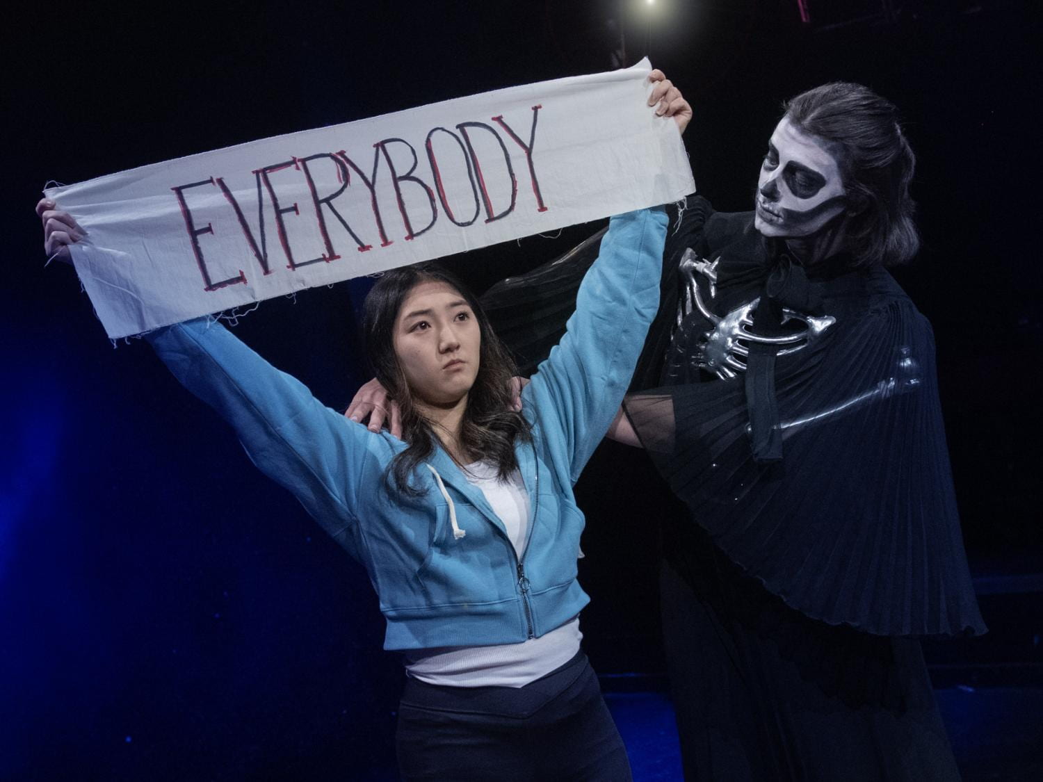Asian young woman holds a sign marked "Everybody," while a young woman dressed as a skeleton peers over her shoulder.