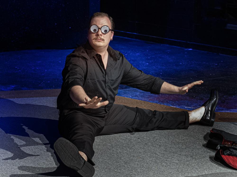 White man with mustache and googly-eyed glasses sits with legs splayed out on the floor and arms reaching out on stage.