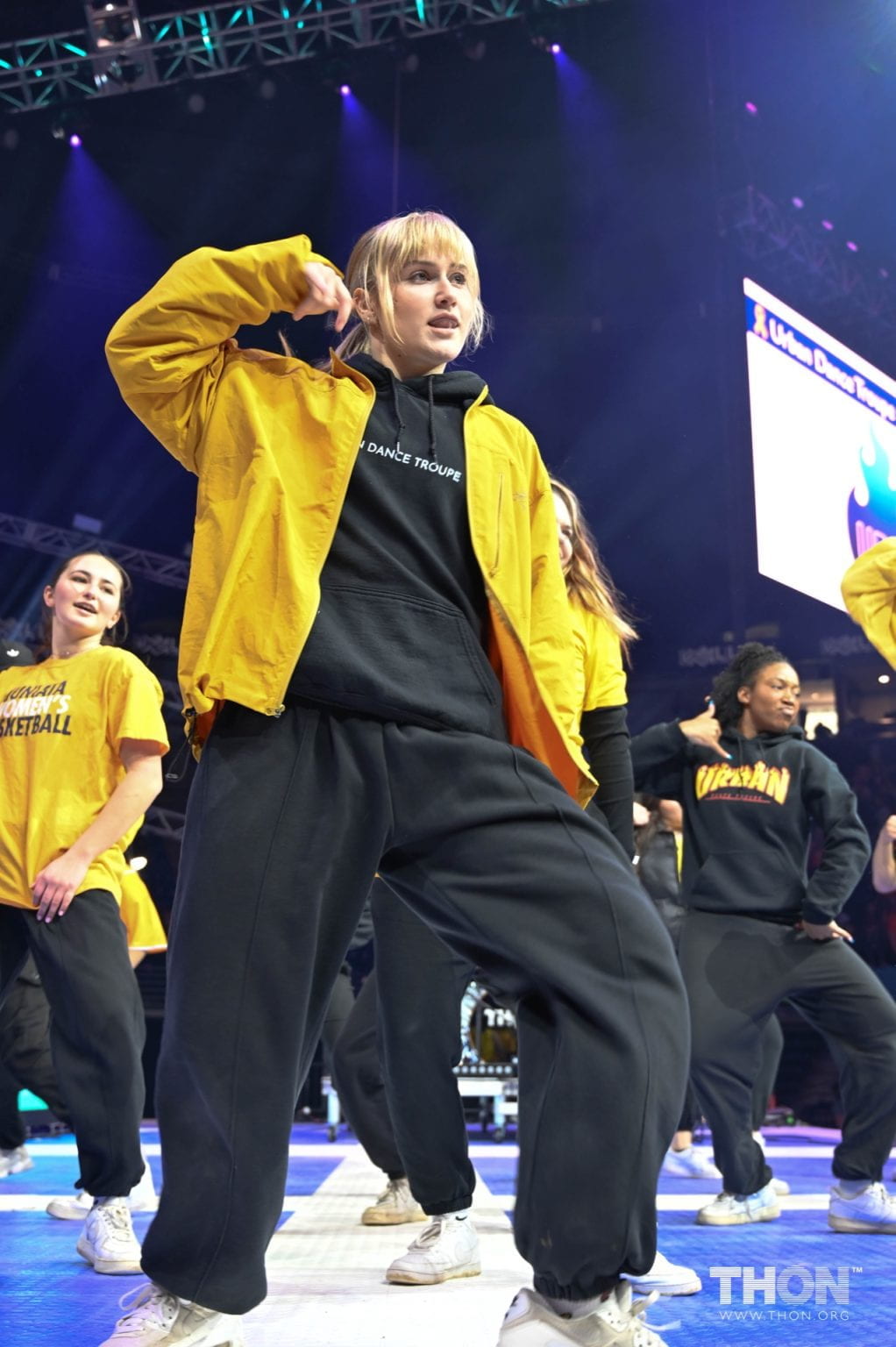 woman wearing black clothes with a yellow jacket poses with hand behind head on stage