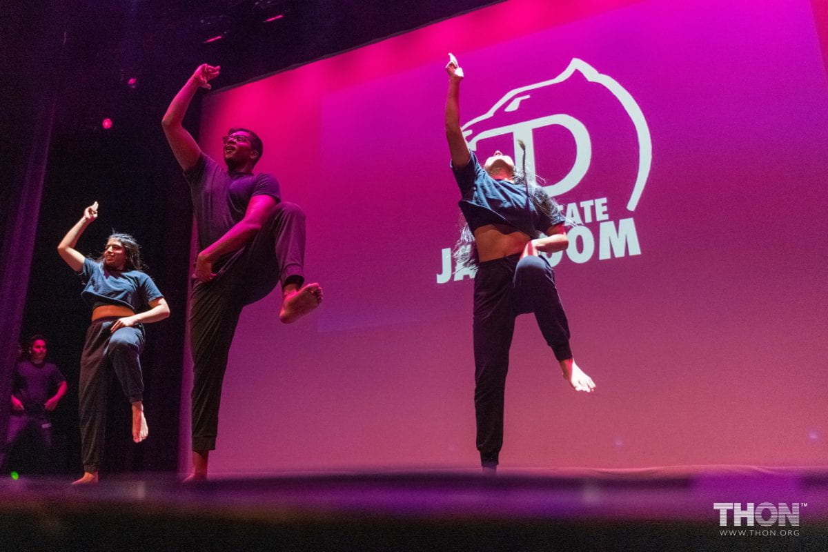 three students stand on pink backlit stage with their right arms in the air and left leg bent up.