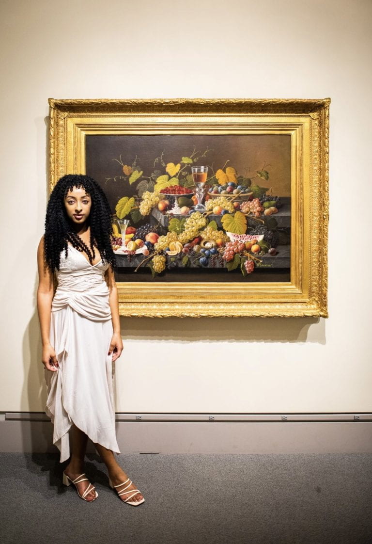 A young woman with dark curly brown hair wearing a beige, flowy dress and standing next to a framed painting of grapes.