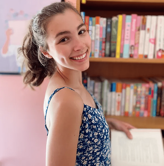 young woman in blue dress smiles over her shoulder in front of a bookcase