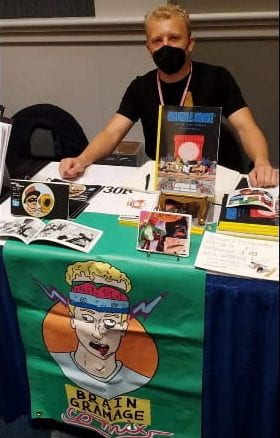 man wearing black mask sits behind table filled with comic books